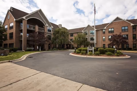 Front exterior of a three-story brick senior living building with a circular driveway, flagpole, and landscaped entrance.