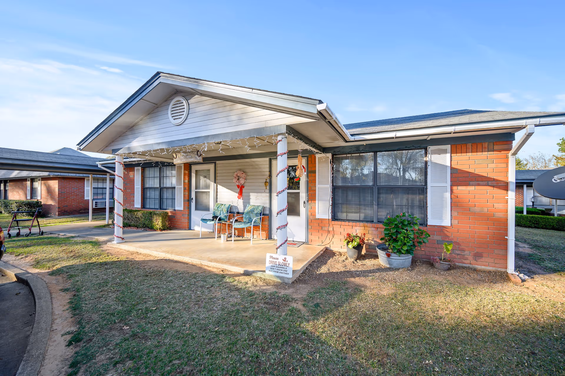 Single-story brick and siding building with a covered front porch, potted plants, holiday decorations, and two chairs.