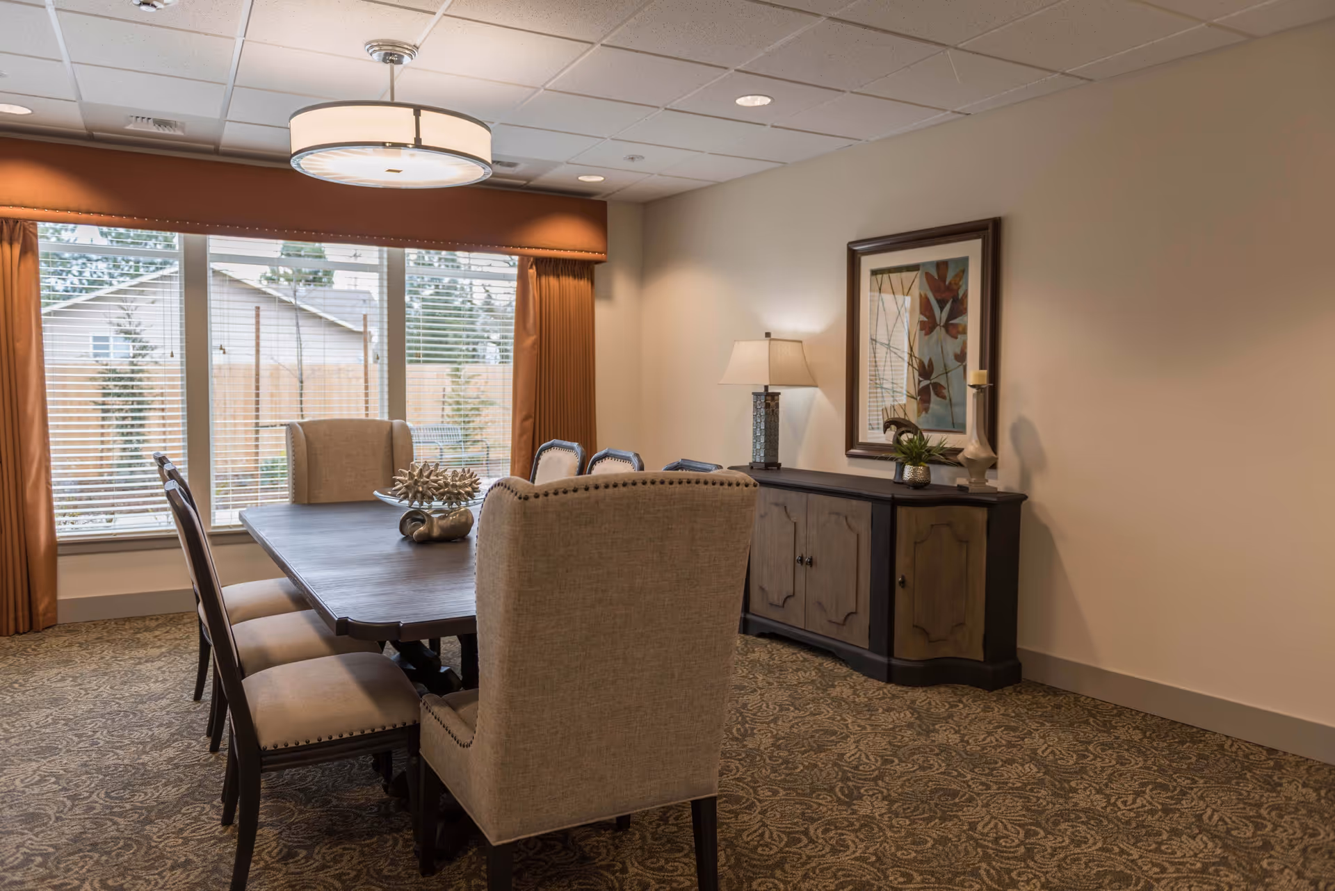 A dining room with a large rectangular wooden table surrounded by eight upholstered chairs. The room has a patterned carpet, a sideboard with a lamp, decorative items, and a framed artwork on the wall. Large windows with blinds and rust-colored curtains allow natural light to enter.