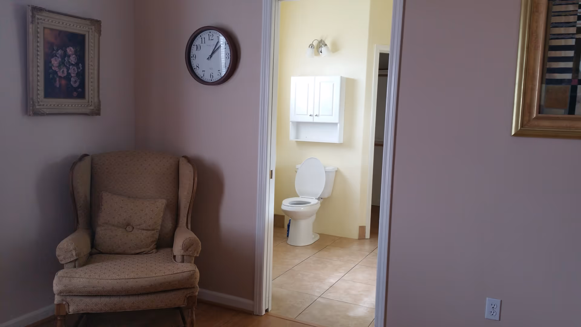 View of a beige upholstered armchair with a matching cushion placed against a light purple wall. Above the chair is a framed floral painting. To the right, an open doorway reveals a bathroom with a white toilet, a white cabinet mounted on a pale yellow wall, and a wall light fixture above the cabinet. A round wall clock is mounted on the wall between the chair and the doorway.