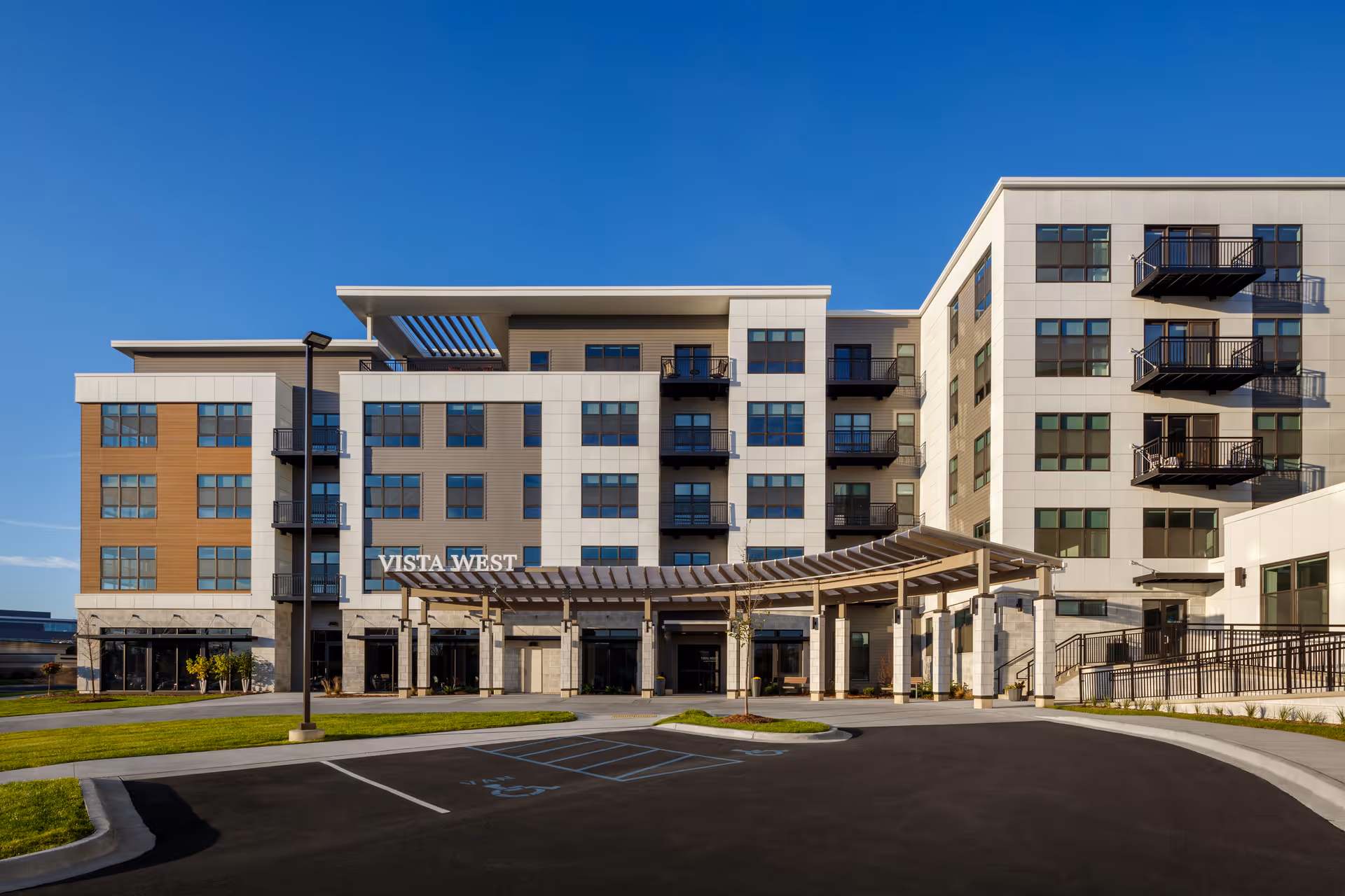 Exterior view of a modern multi-story senior living facility named Vista West with balconies, large windows, a covered entrance, and a parking area with handicap spaces in front.