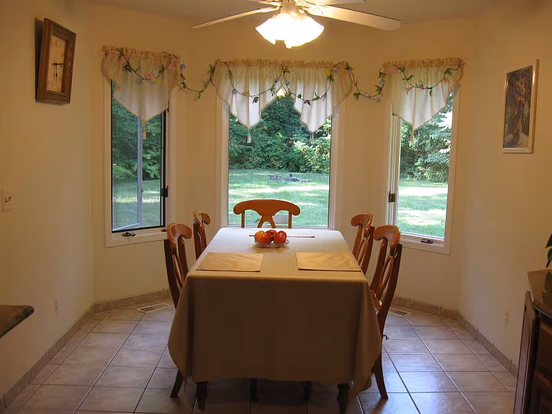 A dining room with a rectangular table covered with a beige tablecloth, surrounded by six wooden chairs. The table has placemats and a bowl of fruit in the center. Three large windows with decorative valances provide a view of a green outdoor area with trees and grass. The room has tiled flooring and a ceiling fan with lights above the table.