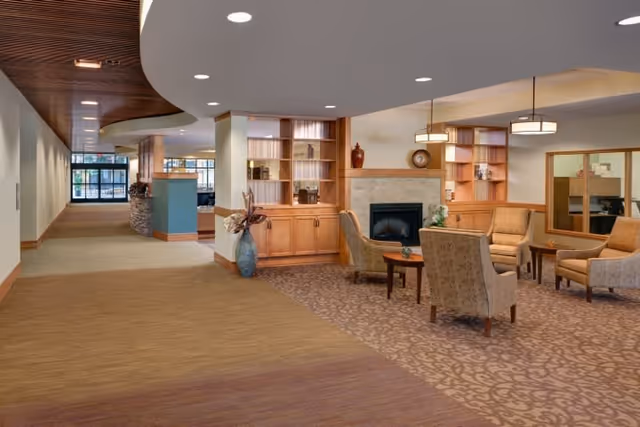 A spacious interior common area with a seating arrangement of four upholstered armchairs around a small wooden table, situated on a patterned carpet. Behind the seating area is a fireplace with built-in wooden shelves and cabinets on either side, decorated with vases and books. The ceiling features recessed lighting and hanging pendant lights. The floor transitions from carpet to wood and then to a lighter carpet leading to a hallway with large windows at the end.