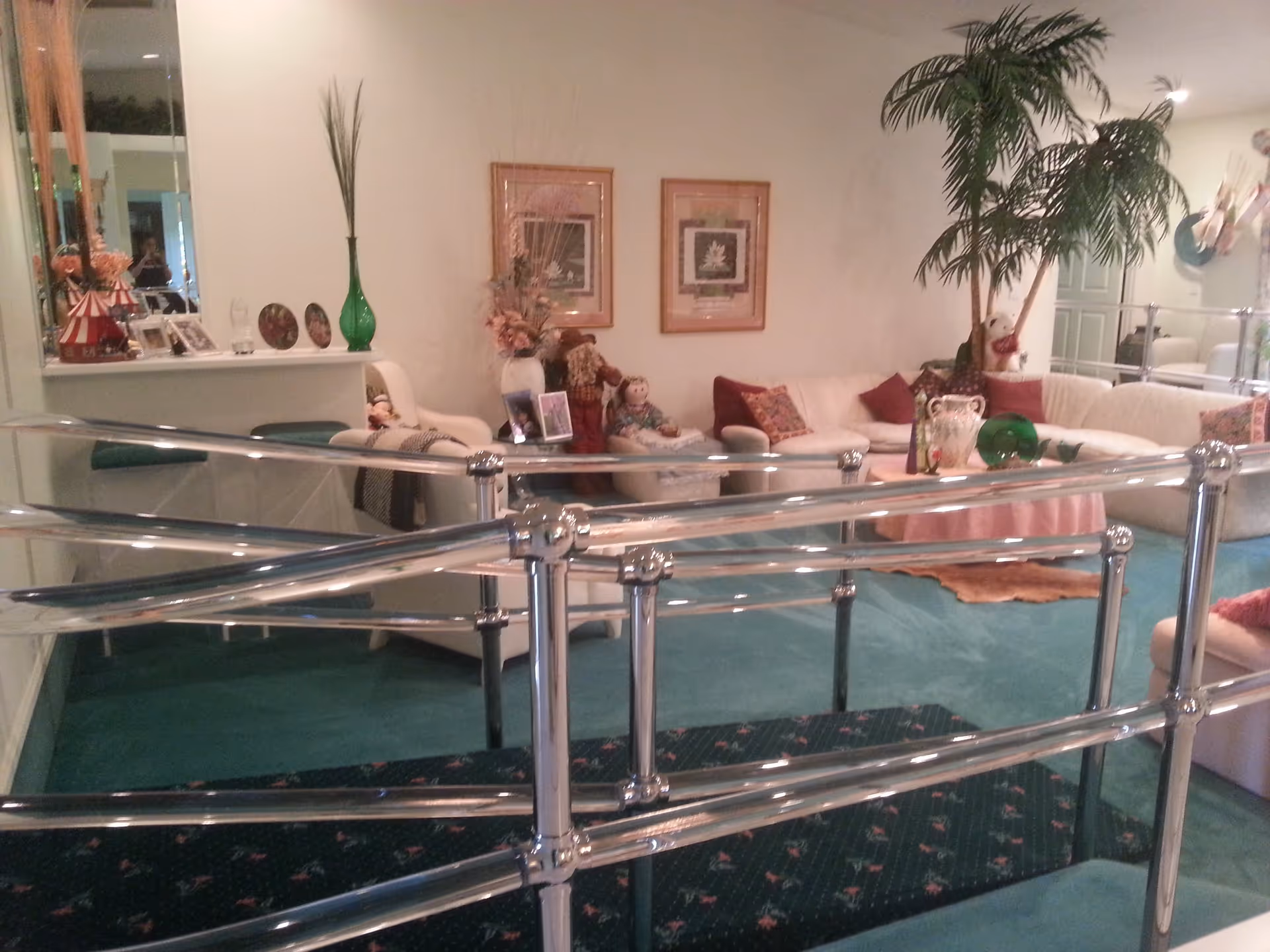 Interior view of a living room with teal carpet and a metal handrail ramp in the foreground. The room features a white sectional sofa with red and patterned pillows, a round table with decorative items, two framed pictures on the wall, a large potted plant, and various decorative dolls and photo frames on a shelf and side table.
