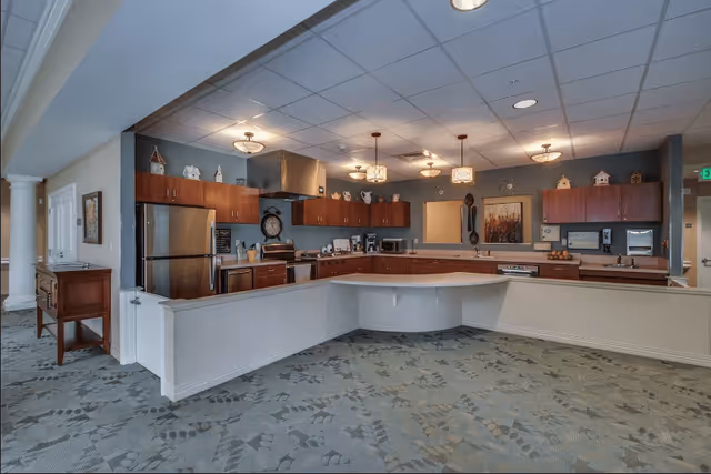 A spacious kitchen area in a senior living facility with wooden cabinets, stainless steel refrigerator, stove, and dishwasher. The kitchen has a large counter with a curved section, decorative items on top of the cabinets, and multiple ceiling lights. The floor is carpeted with a patterned design.