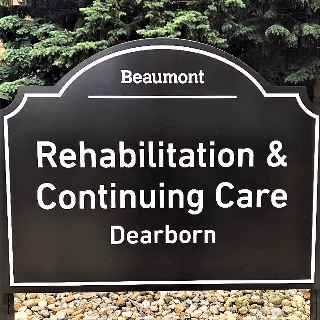 A black sign with white text that reads 'Beaumont Rehabilitation & Continuing Care Dearborn' placed outdoors with green trees in the background and rocks at the base.