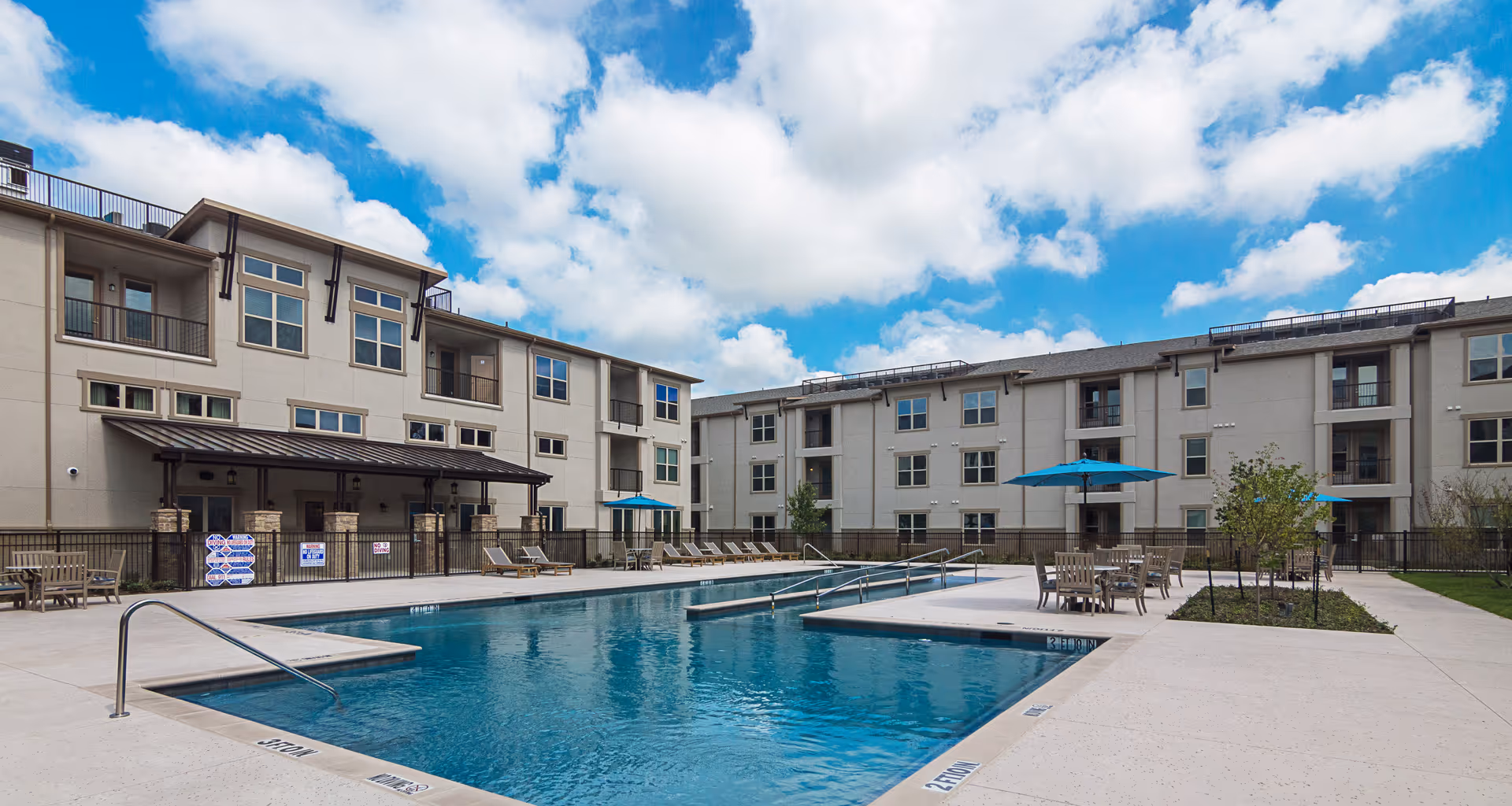 Outdoor courtyard with a swimming pool, lounge chairs, umbrellas, and multi-story apartment buildings under a partly cloudy sky.