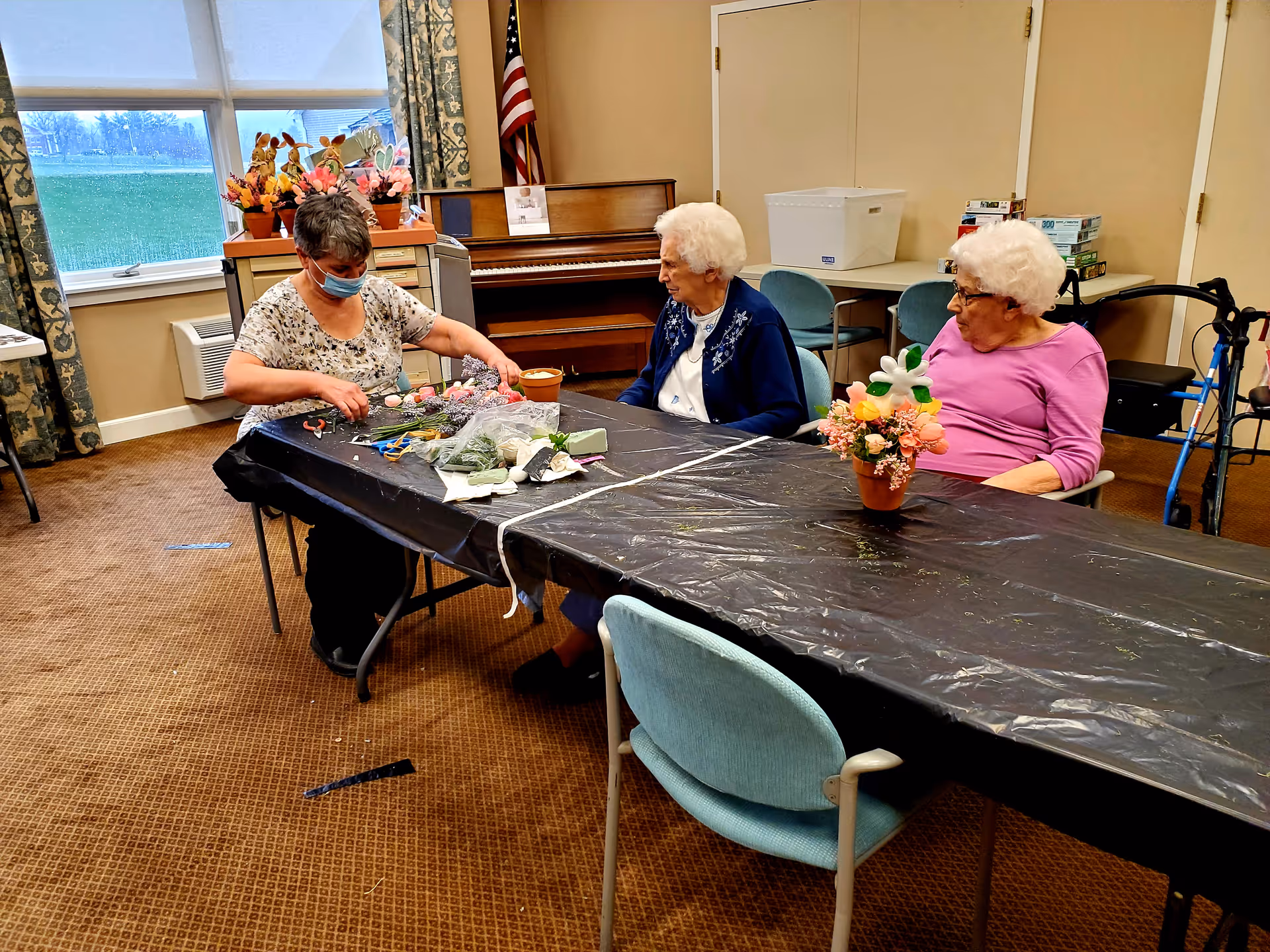 Three elderly women sitting around a table covered with a black plastic tablecloth in a community room. One woman is wearing a face mask and arranging flowers, while the other two women watch. There is a piano, an American flag, and windows with floral curtains in the background. The room has carpeted flooring and several chairs.