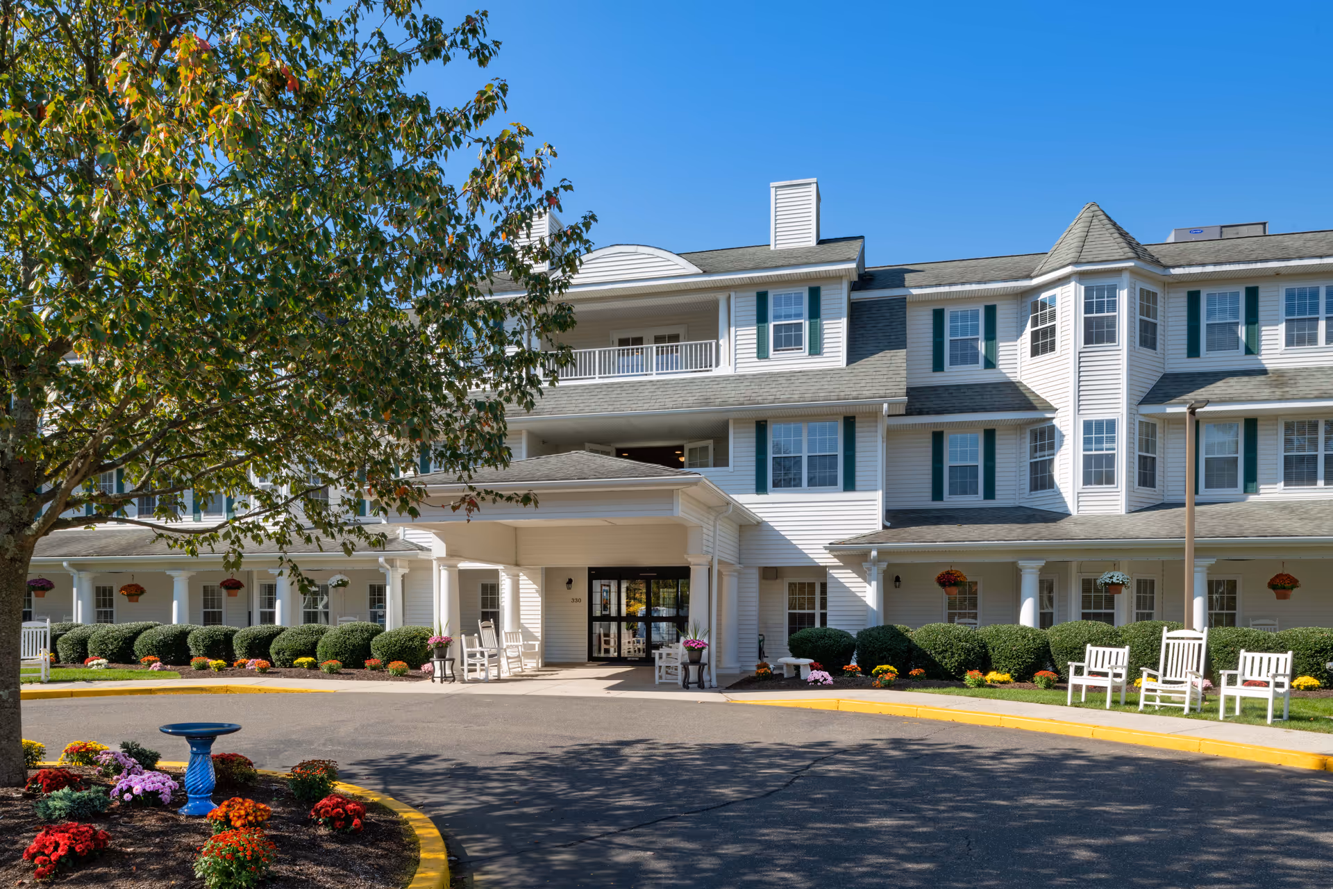 Exterior view of a senior living facility building with white siding and green shutters. The entrance has a covered porch with rocking chairs and potted plants. There are well-maintained bushes, colorful flowers, and a tree in the foreground under a clear blue sky.