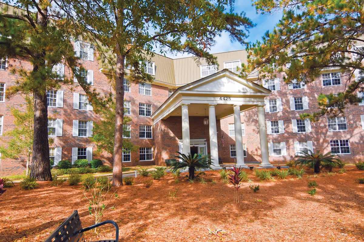 Exterior view of a multi-story brick building with white window shutters and a covered entrance supported by four white columns. The building number 4425 is displayed above the entrance. The foreground features landscaped grounds with pine trees, small plants, and a bench.