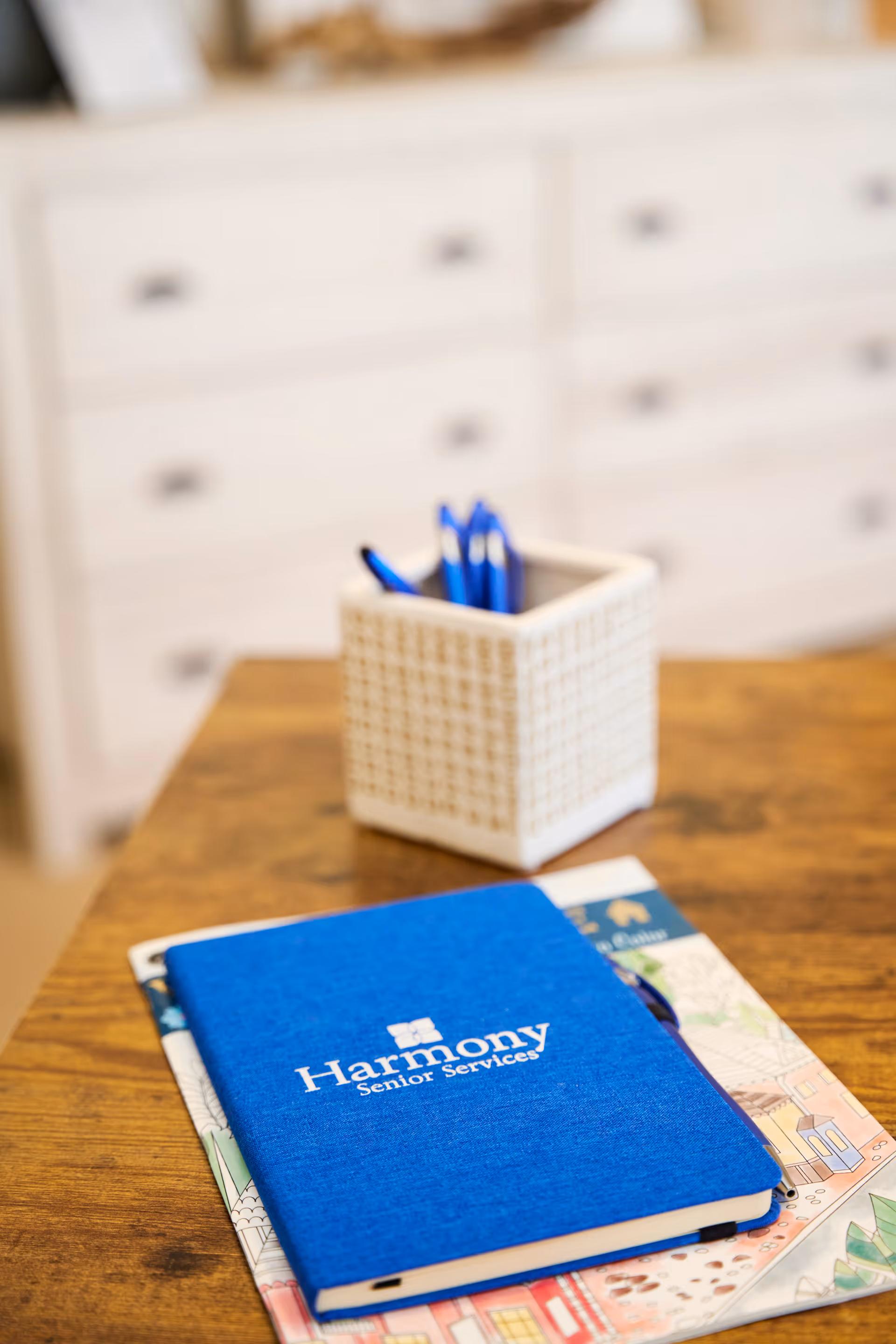 A blue notebook stamped 'Harmony Senior Services' sits on a wooden table beside a white pen holder, with a blurred dresser in the background.