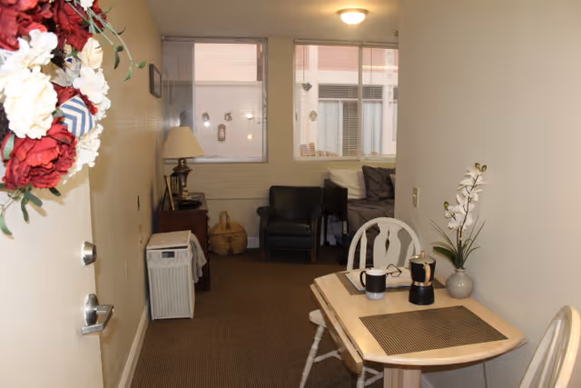 View of a small living area in a senior living facility with a wooden table and two chairs in the foreground. On the table are two mugs, a small coffee maker, a pair of glasses, and a vase with white flowers. In the background, there is a black armchair, a side table with a lamp, a basket, and a couch with cushions. The room has beige walls and carpeted floor, with two windows covered by blinds.
