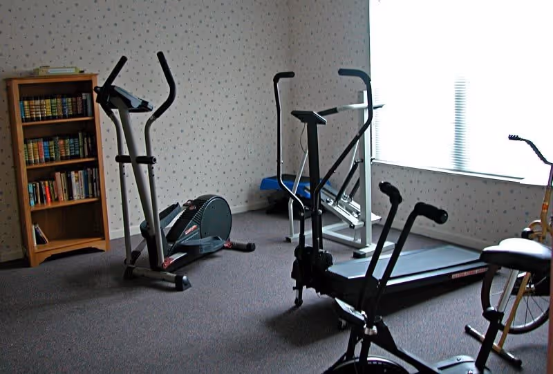 A small exercise room with various fitness equipment including an elliptical machine, a treadmill, and a stationary bike. There is a wooden bookshelf filled with books against the wall, and a large window letting in natural light. The walls have a light-colored wallpaper with a subtle pattern.