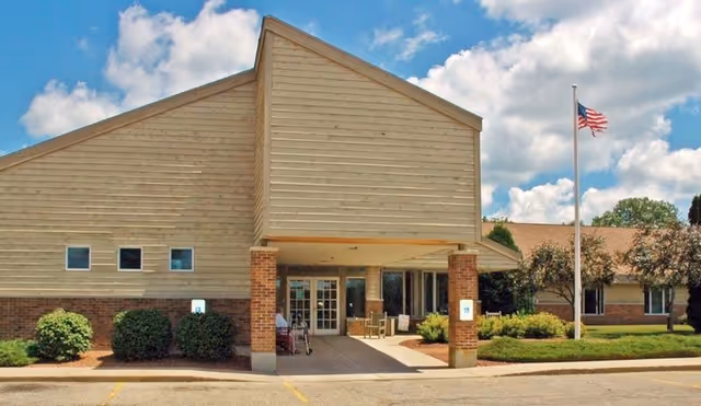 Front entrance of a single-story senior living building with a covered drop-off supported by brick pillars, landscaped shrubs, and an American flag on a flagpole.