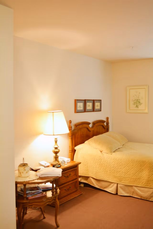 A cozy bedroom with a wooden headboard bed covered in a yellow quilt and matching pillows. Next to the bed is a wooden nightstand with a brass lamp, a clock, and some small items. There is also a small wooden side table with books and a decorative item. The walls are light-colored with framed artwork hanging above the bed and on the adjacent wall.