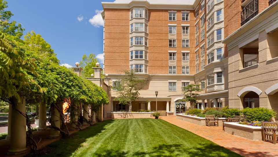 Brick multi-story senior living building with a green lawn courtyard, pergola-covered walkway and benches.