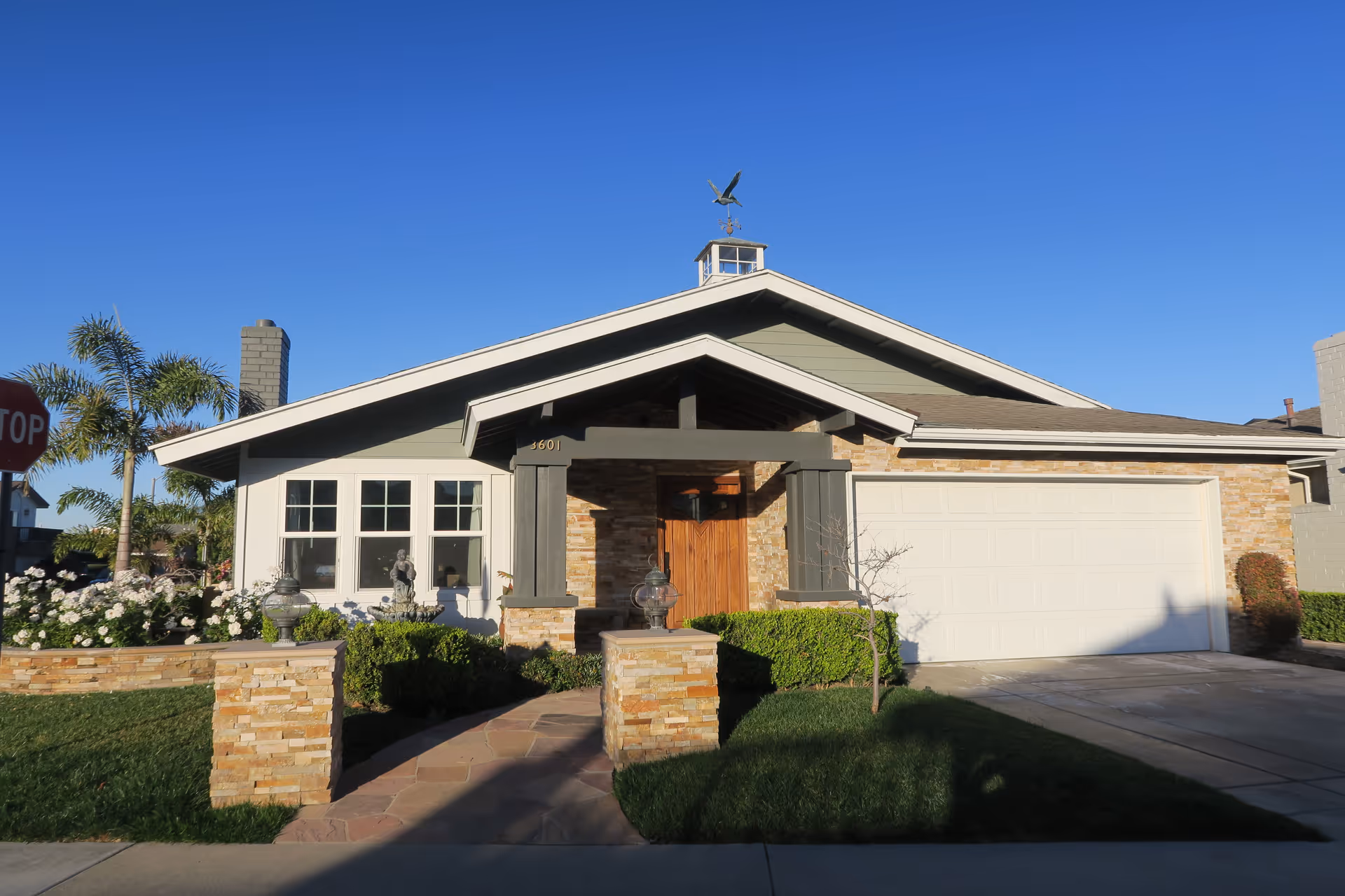 Front exterior view of a single-story house with a stone and wood facade, a large garage door, a pathway leading to the wooden front door, and well-maintained landscaping including bushes and flowers under a clear blue sky.
