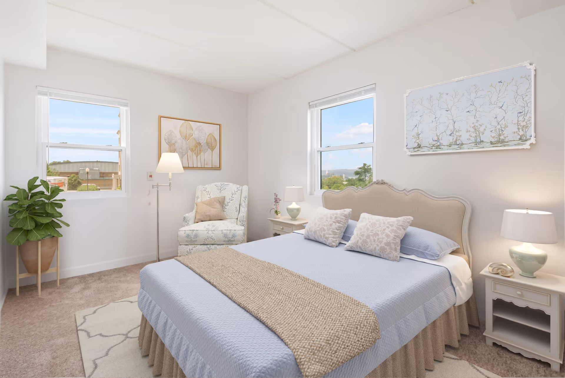 A bright and cozy bedroom in Drum Hill Senior Living Community featuring a large bed with light blue bedding and two decorative pillows. The room has two windows letting in natural light, a patterned armchair with a beige pillow, a floor lamp, two nightstands with lamps, a potted plant, and framed artwork on the walls.