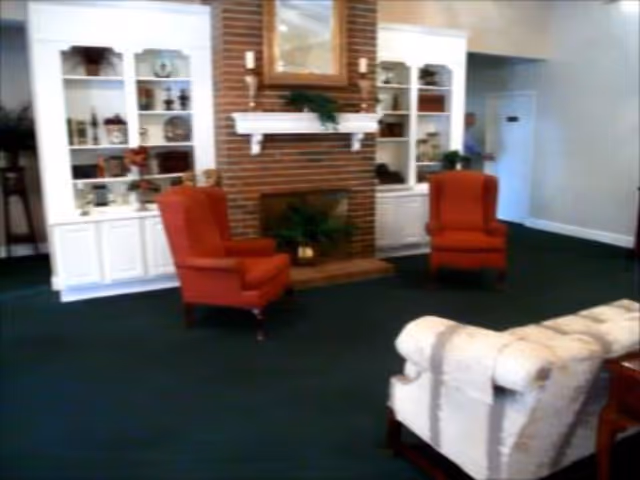 A cozy living room area with a brick fireplace in the center, flanked by two white built-in shelves filled with decorative items. There are two red upholstered armchairs facing the fireplace and a light-colored upholstered sofa in the foreground. The room has dark green carpet and light-colored walls.
