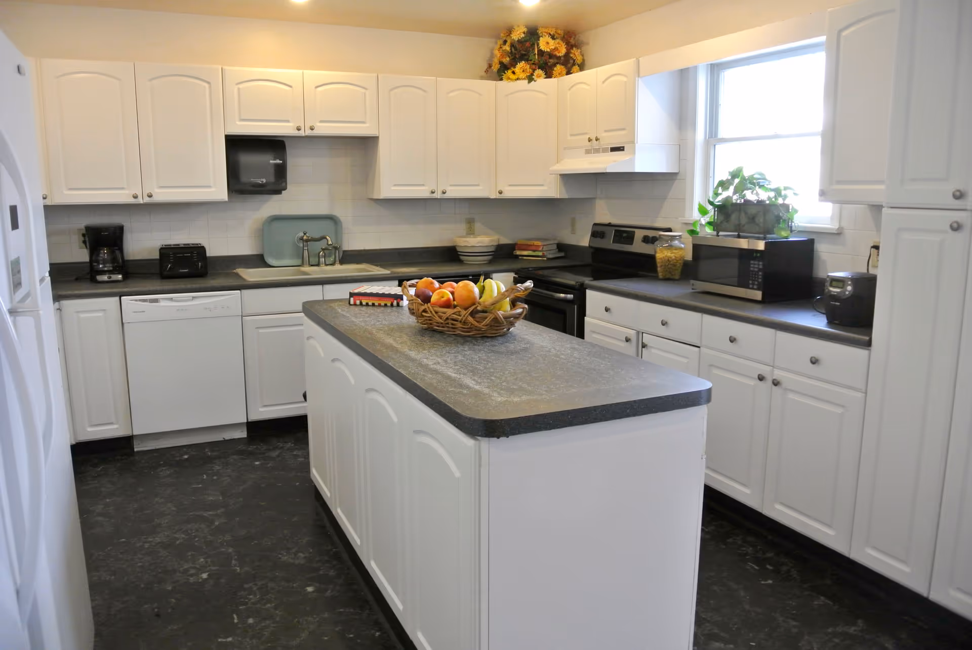 A clean and bright kitchen with white cabinets and black countertops. The kitchen features a central island with a basket of fruit on top, a dishwasher, a coffee maker, a toaster, a stove with an oven, a microwave, and a window with a potted plant on the counter. There is a bouquet of yellow flowers on top of the cabinets.