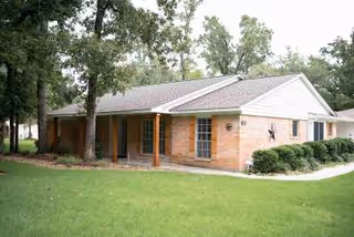 Single-story brick building with a white gabled roof surrounded by grass and trees, featuring a covered porch supported by wooden posts.