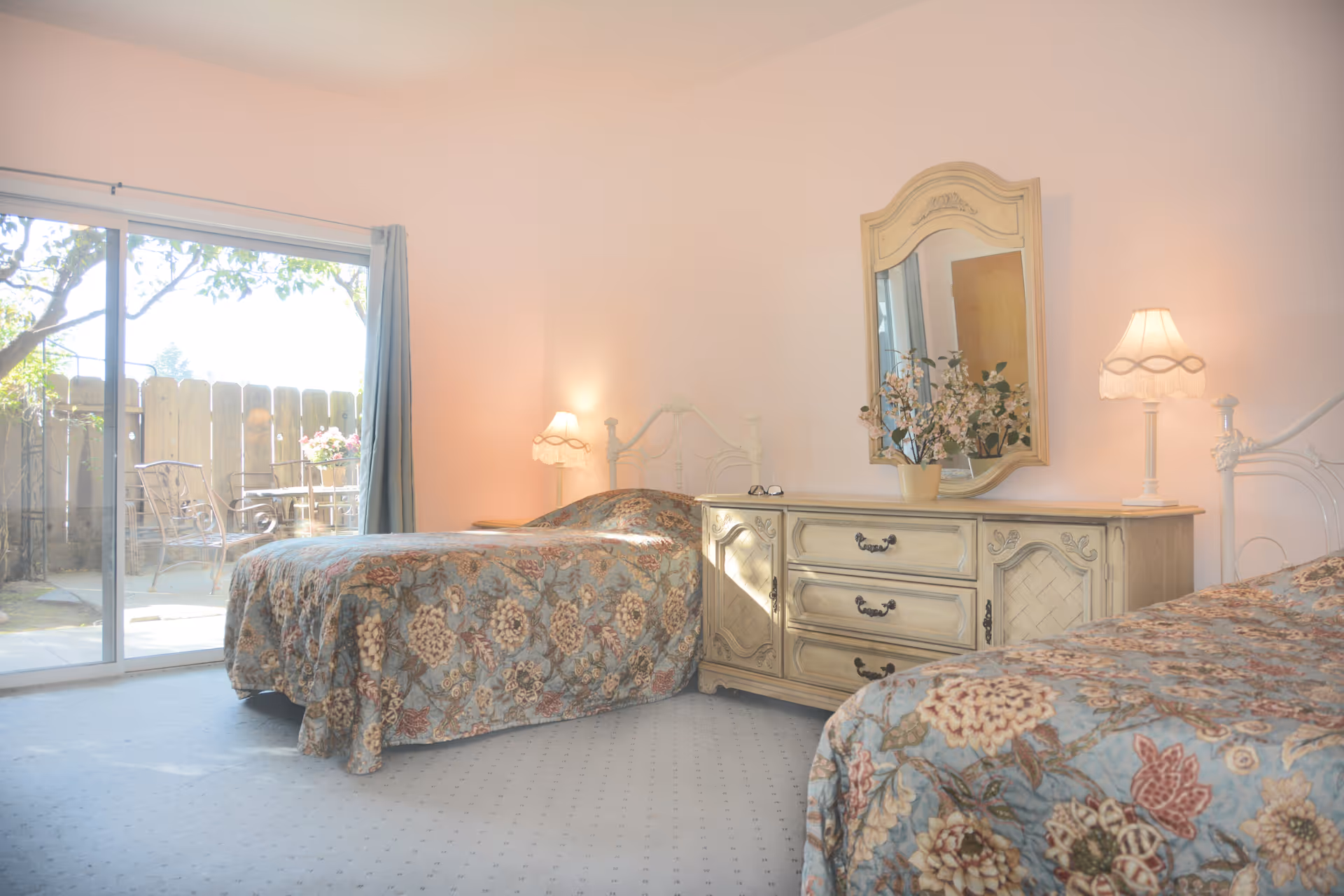 A bright bedroom with two twin beds covered in floral patterned bedspreads. Between the beds is a vintage-style wooden dresser with a large mirror and a vase with flowers on top. Two lamps with fringed shades are on either side of the dresser. A sliding glass door leads to an outdoor patio with metal chairs and a table, surrounded by a wooden fence and greenery.