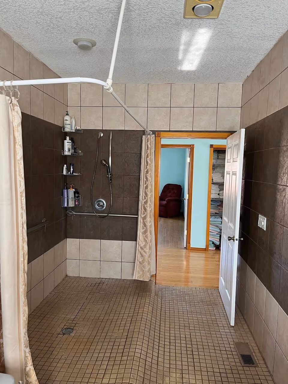 A tiled bathroom with a walk-in shower area featuring beige and dark brown tiles. The shower has a handheld showerhead, grab bars, and a corner shelf with various toiletries. A shower curtain is partially drawn. The bathroom door is open, revealing a hallway with wooden flooring, a room with a red armchair, and a closet with neatly stacked towels.