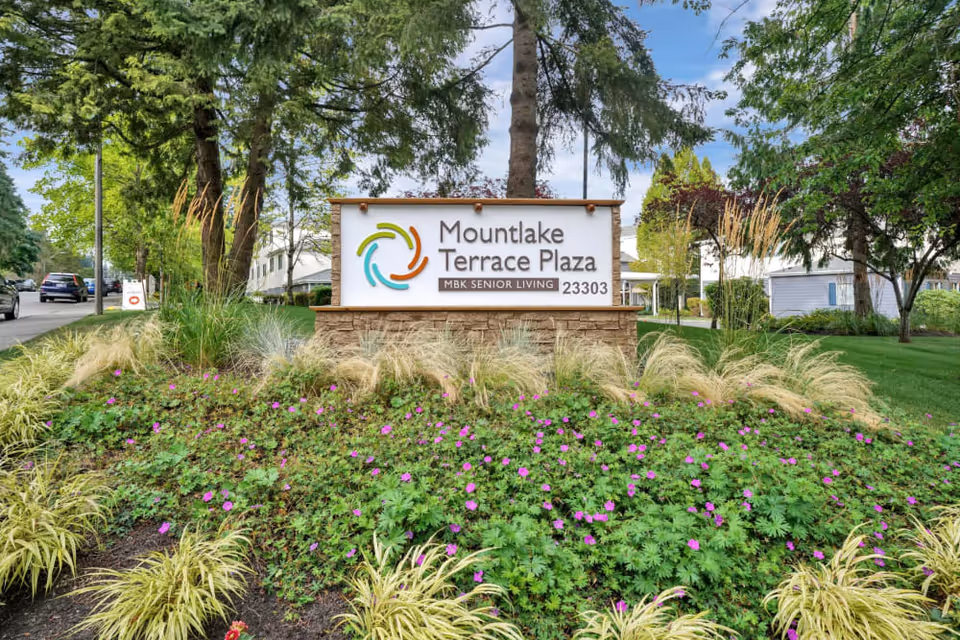 Entrance sign for Mountlake Terrace Plaza surrounded by landscaping, flowering plants, and trees.