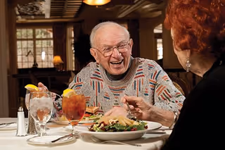 An elderly man and woman sitting at a dining table in a warmly lit room, enjoying a meal together. The man is smiling and wearing glasses and a patterned sweater, while the woman is partially visible with red hair. The table is set with plates of food, glasses of iced tea with lemon, and silverware.