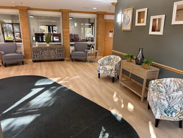 A bright and welcoming interior common area with light wood flooring and a large dark rug. The room features several upholstered chairs, including two with a floral pattern and two solid gray armchairs. There is a wooden console table with decorative plants and vases, and framed artwork on the gray wall. Large windows and glass panels allow natural light to fill the space.