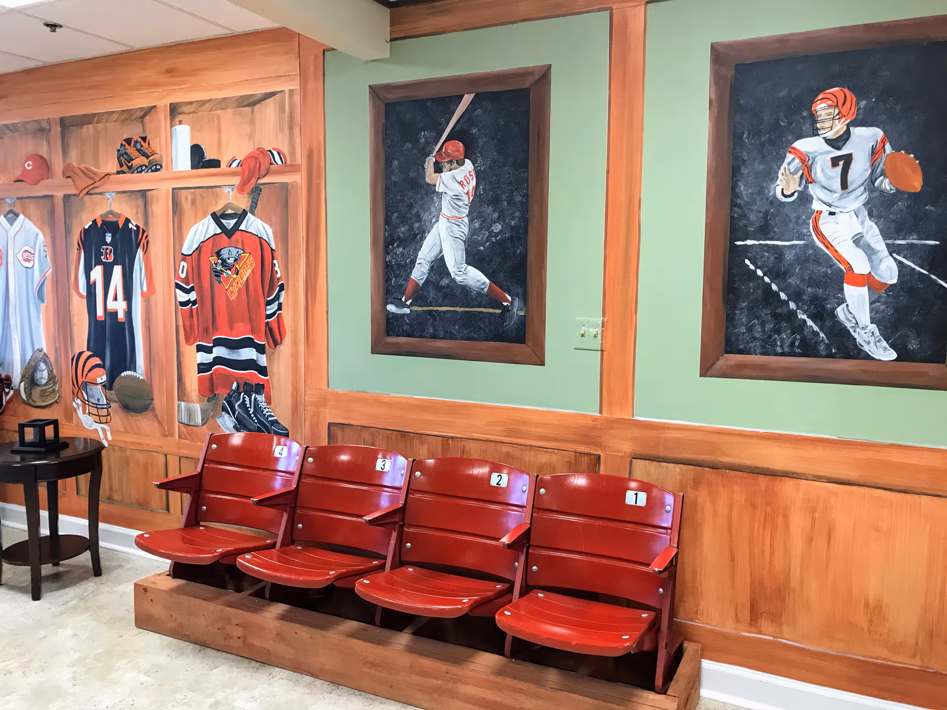 A seating area with four red stadium-style chairs numbered 1 to 4 mounted on a wooden base. The wall behind features sports-themed murals including baseball and football players, as well as painted sports jerseys, helmets, and equipment on wooden panels.