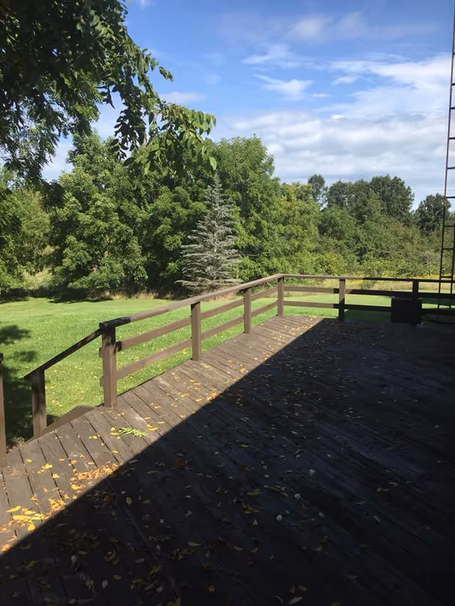 A wooden deck with a railing overlooking a green grassy area and dense trees under a partly cloudy blue sky.