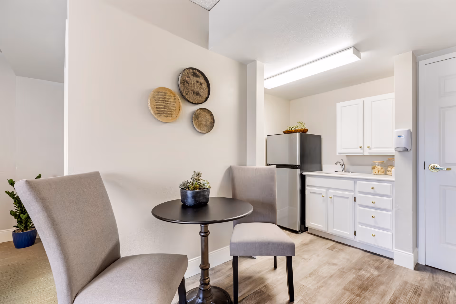 Small kitchenette area with a black round table and two beige upholstered chairs. On the table is a small potted succulent plant. The kitchenette has white cabinets, a stainless steel mini refrigerator, a sink, and two jars on the counter. Three decorative round wall hangings are mounted on the wall above the table. The floor is wood-style laminate, and there is a white door to the right.
