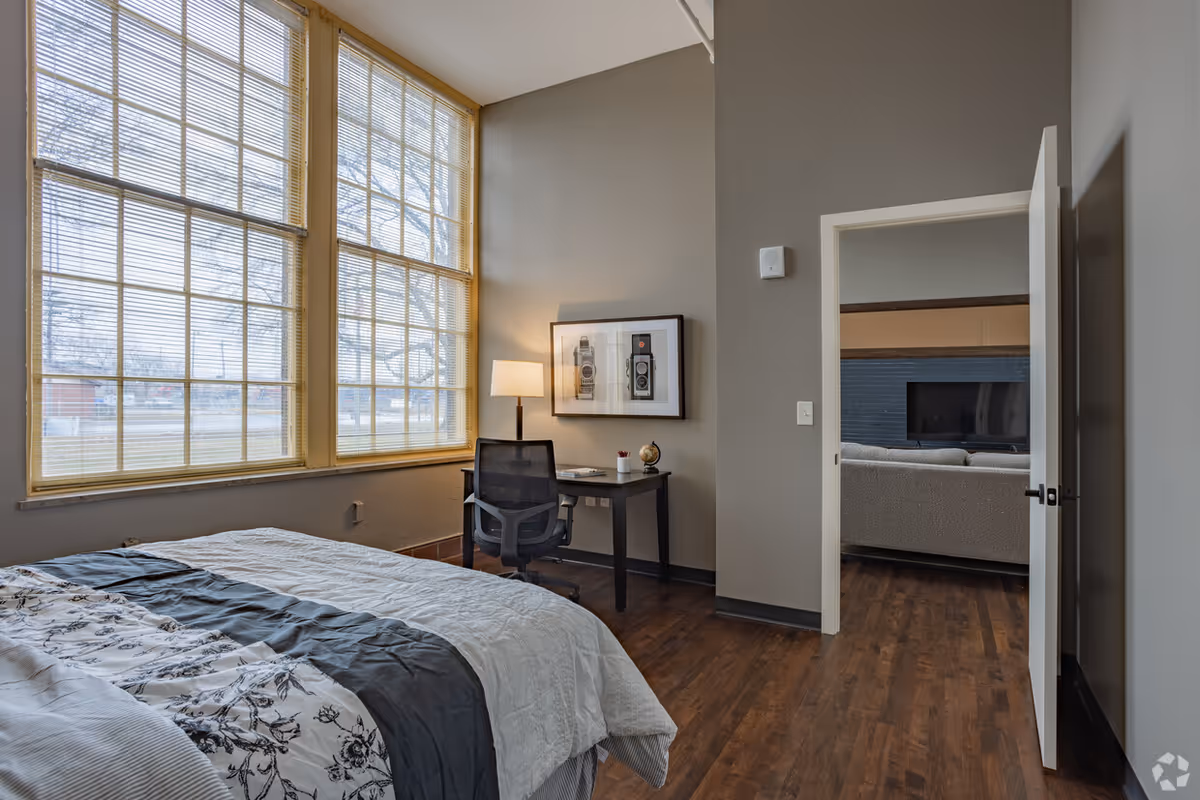 A bedroom with a large window covered by blinds, a bed with a white and gray floral comforter, a small black desk with a lamp, chair, and framed pictures on the wall. The door is open, showing a glimpse of a living room with a TV and sofa.