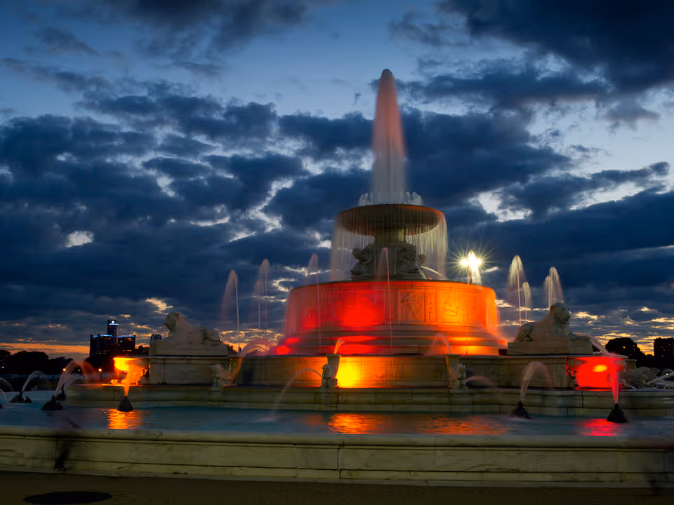 A large illuminated fountain at dusk with water jets spraying upwards, surrounded by sculpted lion statues. The sky is cloudy with a hint of sunset colors in the background.