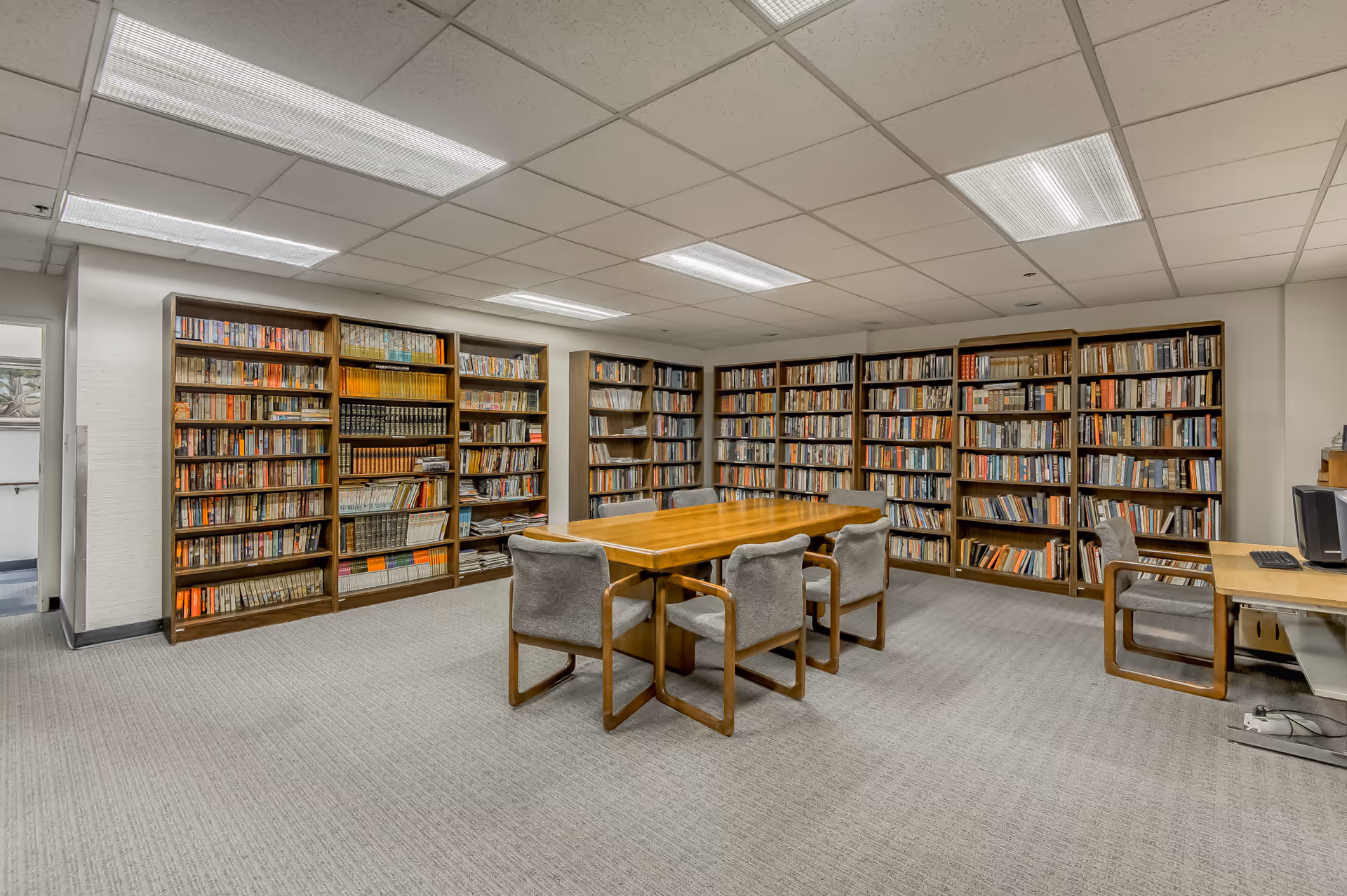 A quiet library room with multiple wooden bookshelves filled with books along the walls. In the center, there is a wooden table surrounded by six cushioned chairs. The room has a carpeted floor and a drop ceiling with fluorescent lights. A computer workstation is visible on the right side of the image.