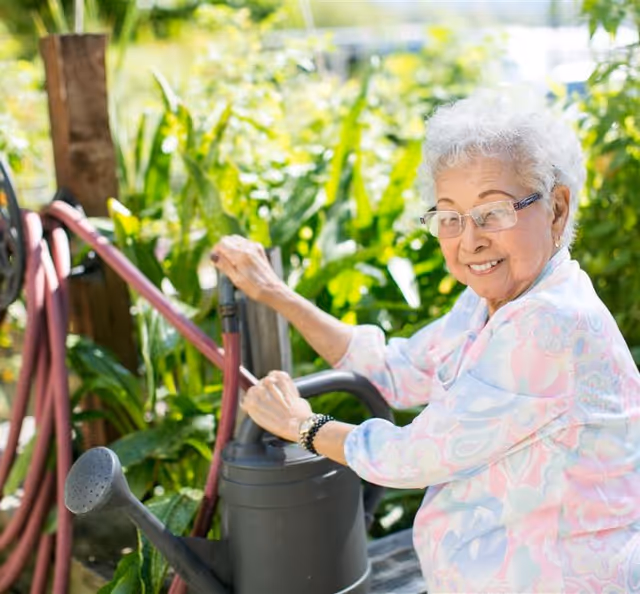 An elderly woman with short gray hair and glasses is smiling while watering plants in a garden using a black watering can. She is wearing a light-colored patterned blouse and is surrounded by lush green foliage.
