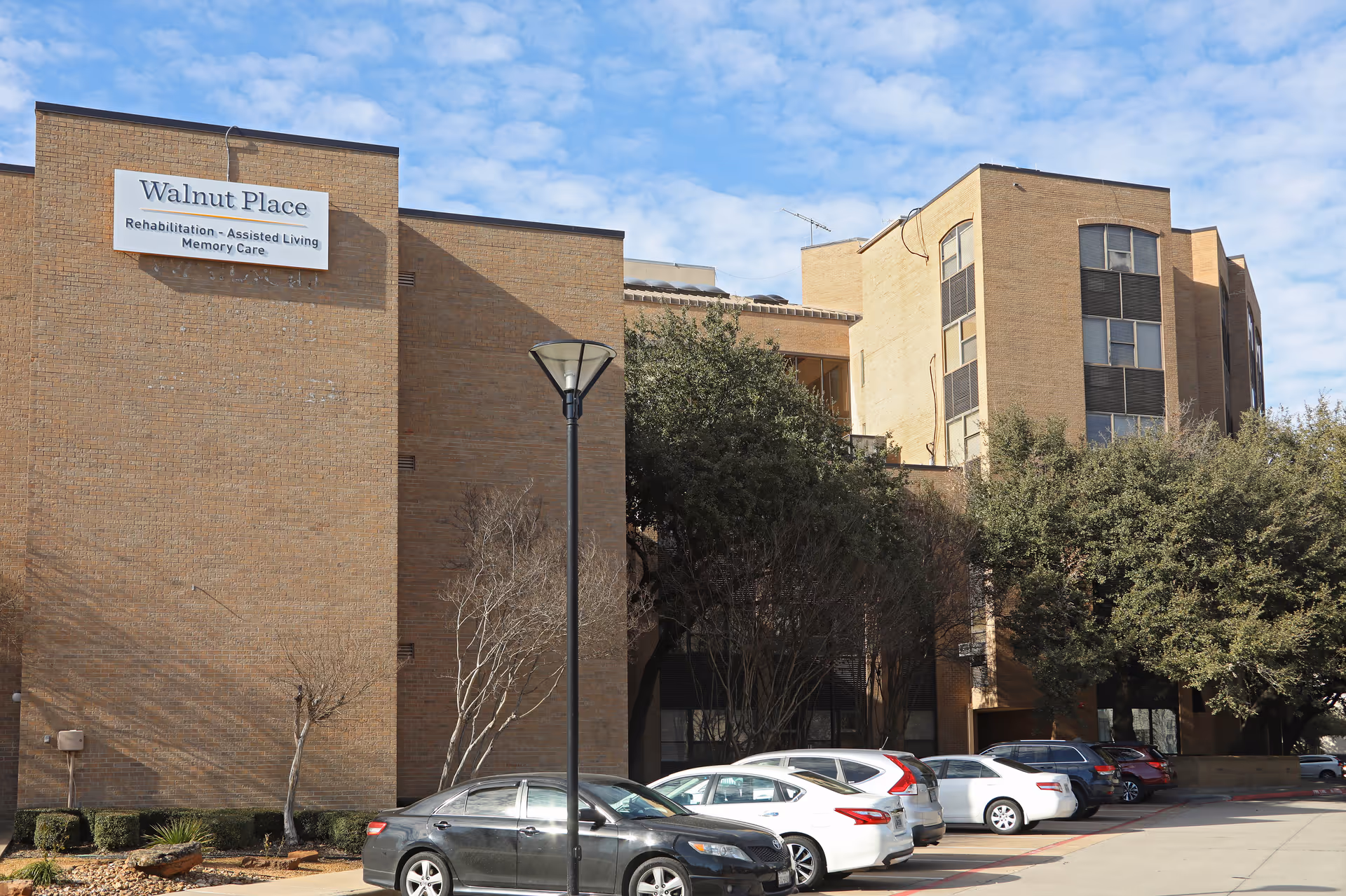 Exterior view of Walnut Place, a multi-story brick building with a sign indicating Rehabilitation, Assisted Living, and Memory Care services. Several cars are parked in front of the building under a partly cloudy sky.