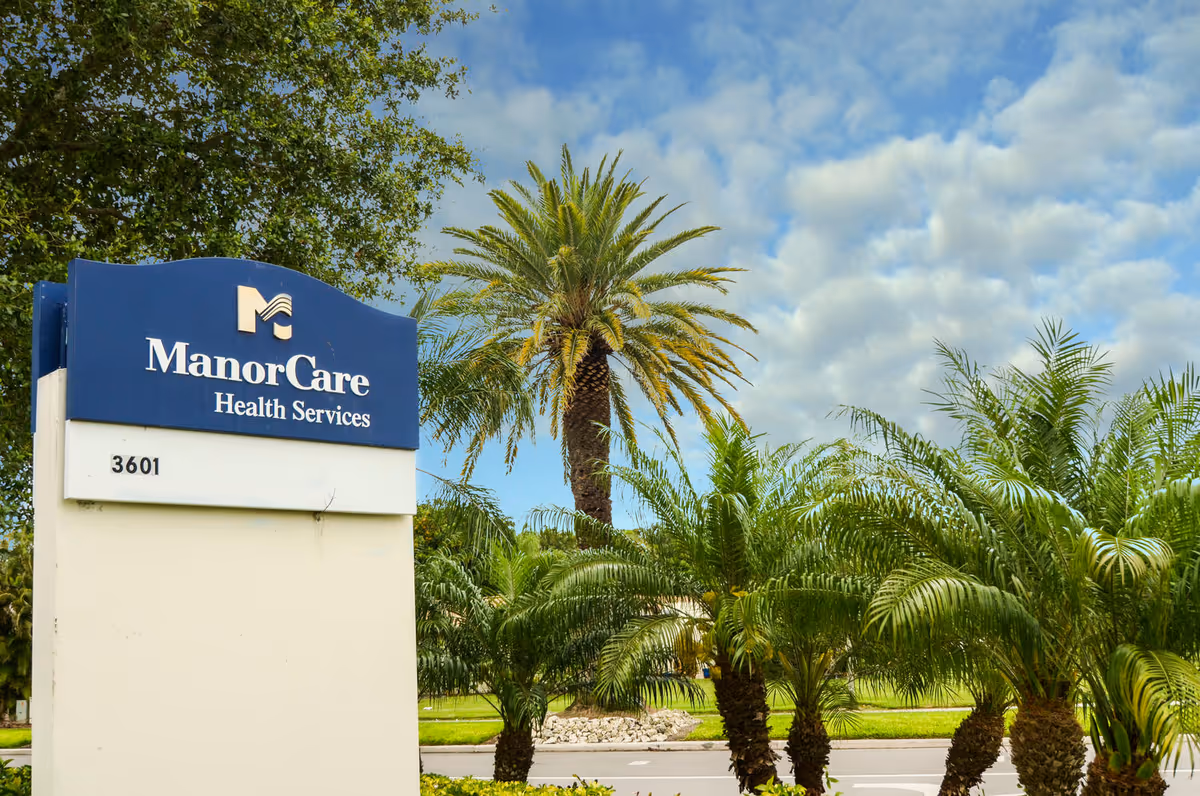 Outdoor view of a ManorCare Health Services sign with the address number 3601, surrounded by palm trees and greenery under a partly cloudy sky.