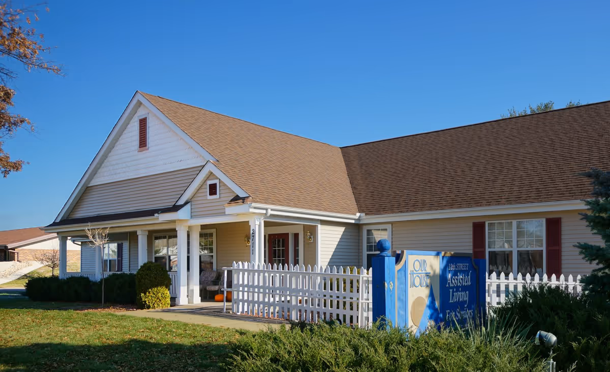 Exterior view of a single-story assisted living facility building with beige siding, a brown shingled roof, white columns, and a white picket fence. There is a blue sign in front that reads 'Our House 12th Street Assisted Living For Seniors'. The sky is clear and blue.