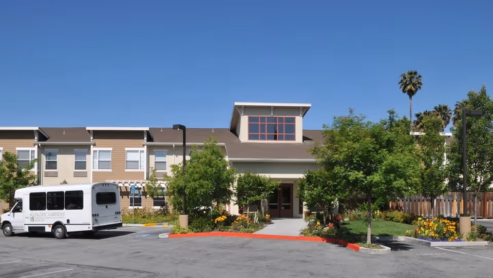 Exterior view of the Pacific Gardens senior living facility building with a white shuttle bus parked in front. The building is two stories with beige and white walls, surrounded by green trees and colorful flowers under a clear blue sky.