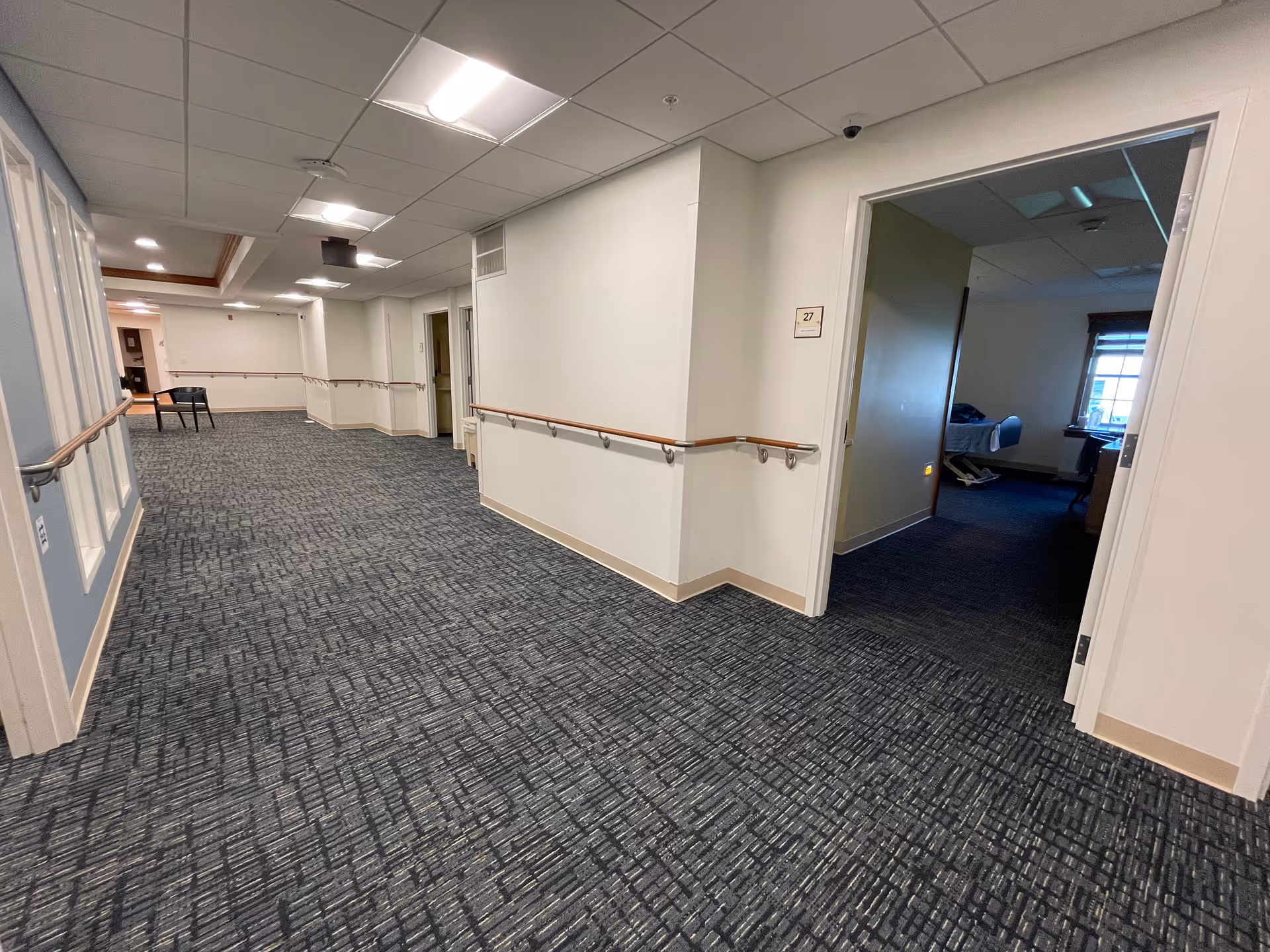 A wide hallway in a senior living facility with blue patterned carpet and white walls. Handrails run along both sides of the hallway. There is an open door on the right side leading to a room with a bed and a window with blinds. The hallway is well-lit with ceiling lights and has a table with chairs at the far end.