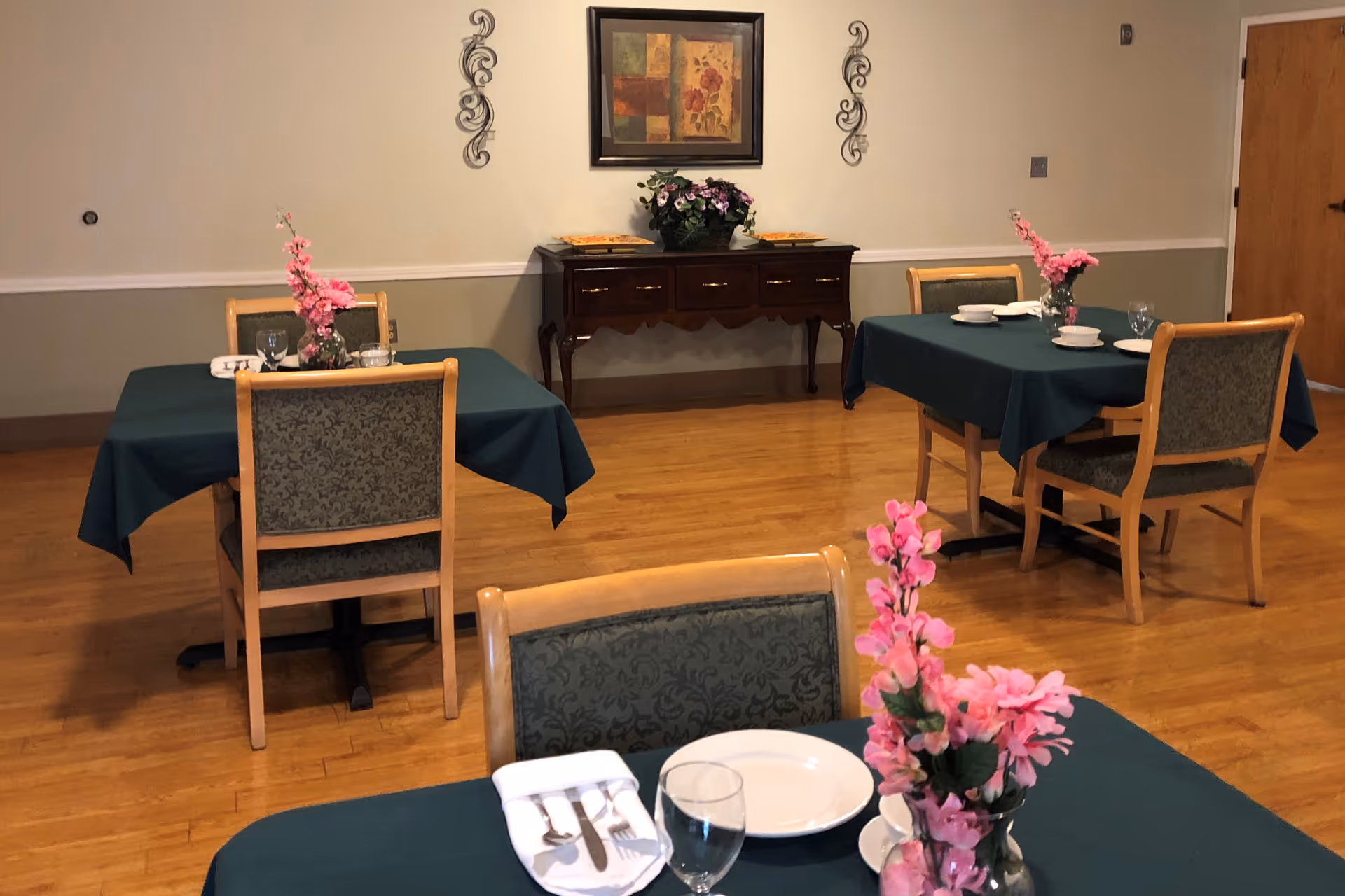 A small dining room with three tables covered in dark green tablecloths, each set with plates, glasses, and silverware. Pink floral centerpieces are placed on each table. In the background, there is a wooden sideboard with a flower arrangement and a framed painting on the wall above it. The room has wooden flooring and neutral-colored walls.