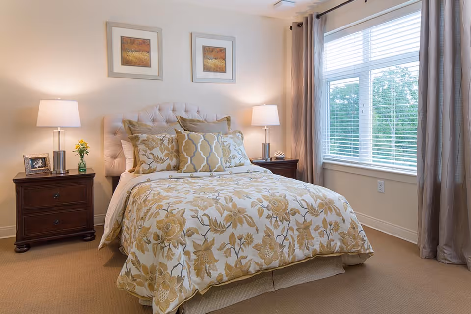 A cozy bedroom with a bed featuring a floral patterned comforter and multiple decorative pillows. There are two wooden nightstands on either side of the bed, each with a lamp. A small vase with yellow flowers and a framed photo are on the left nightstand. The room has beige walls, a large window with blinds and curtains, and two framed artworks hanging above the bed.