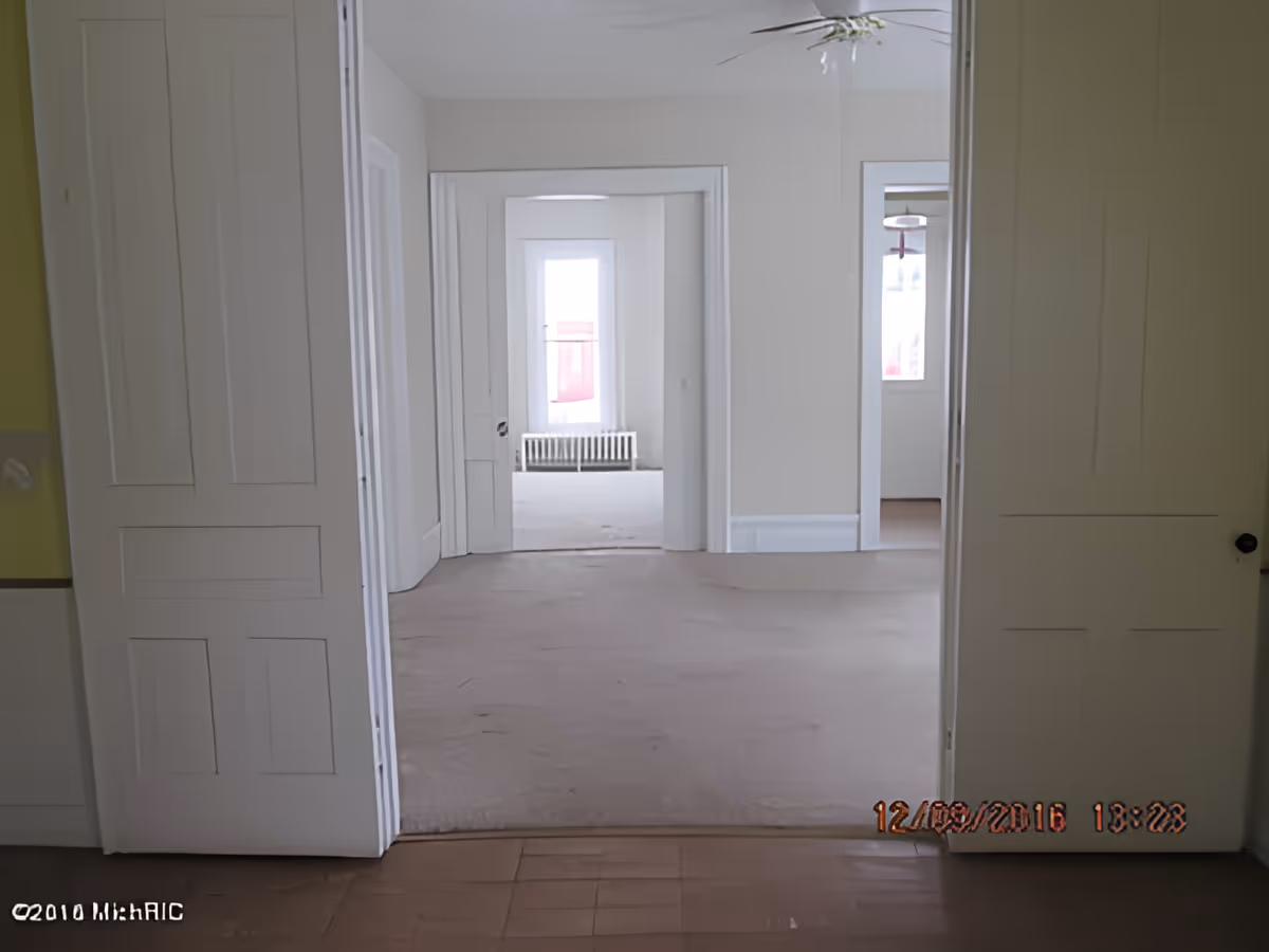 View through an open doorway into an empty room with white walls and a window with a radiator beneath it. The floor appears to be bare and the room is unfurnished. There is a ceiling fan visible in the room on the right.