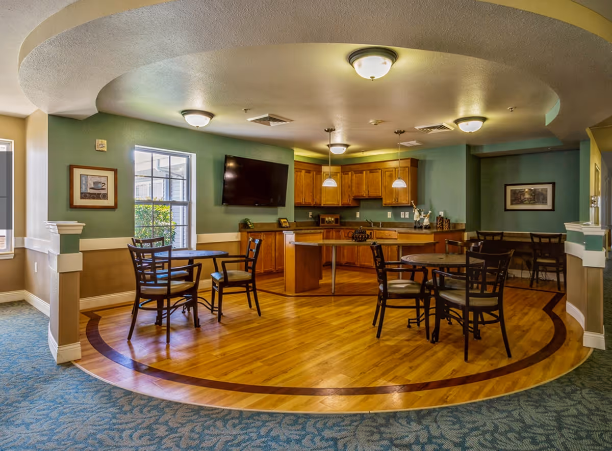 Communal dining area with round tables and chairs on wood flooring in front of a kitchen counter and a wall-mounted TV.