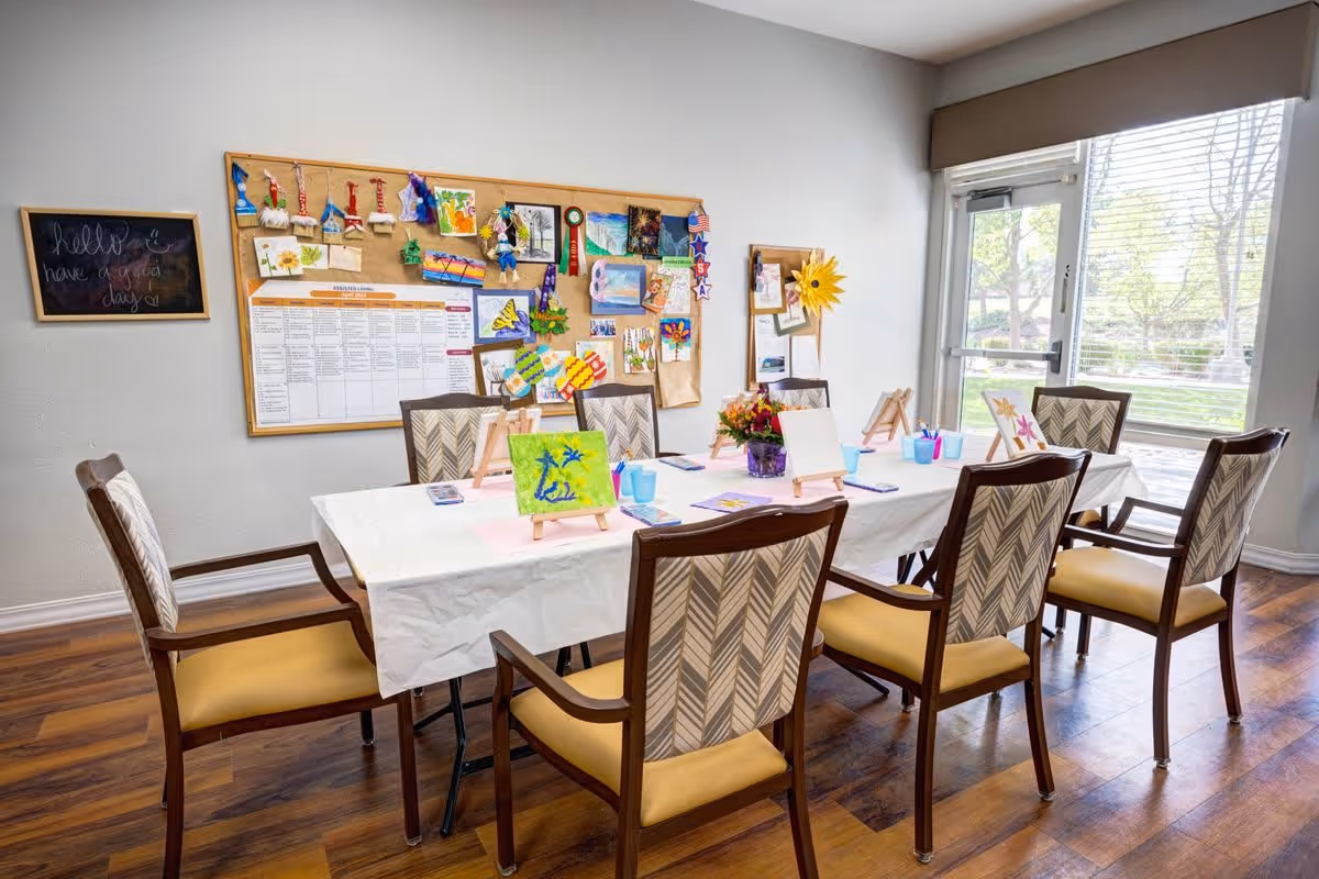 A bright activity room with a rectangular table covered with a white tablecloth, surrounded by eight chairs with patterned backs and yellow seats. The table is set up for painting with small canvases on easels, paint cups, and brushes. On the wall behind the table is a bulletin board displaying various colorful artwork and a schedule, and a small chalkboard with the message 'hello have a good day'. Large windows on the right side let in natural light and show a view of trees outside.