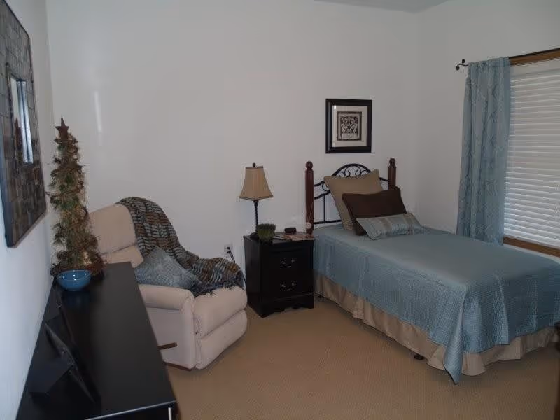 A small bedroom with a single bed covered in a blue bedspread and multiple pillows. Next to the bed is a black nightstand with a lamp, a small plant, and some items on it. There is a beige recliner chair with a blue and brown blanket draped over it. A window with blue curtains and closed blinds is on the right wall. A decorative framed picture hangs above the bed, and a small artificial tree is on a black dresser on the left side of the room.