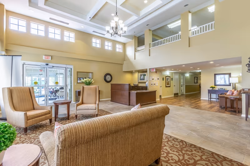 A spacious and well-lit senior living facility lobby with high ceilings and multiple windows. The area features comfortable seating including a sofa and armchairs arranged around small tables, a reception desk, and decorative elements such as a wall clock and framed artwork. The flooring transitions from carpet to tile and wood, and there are exit signs above the doors.