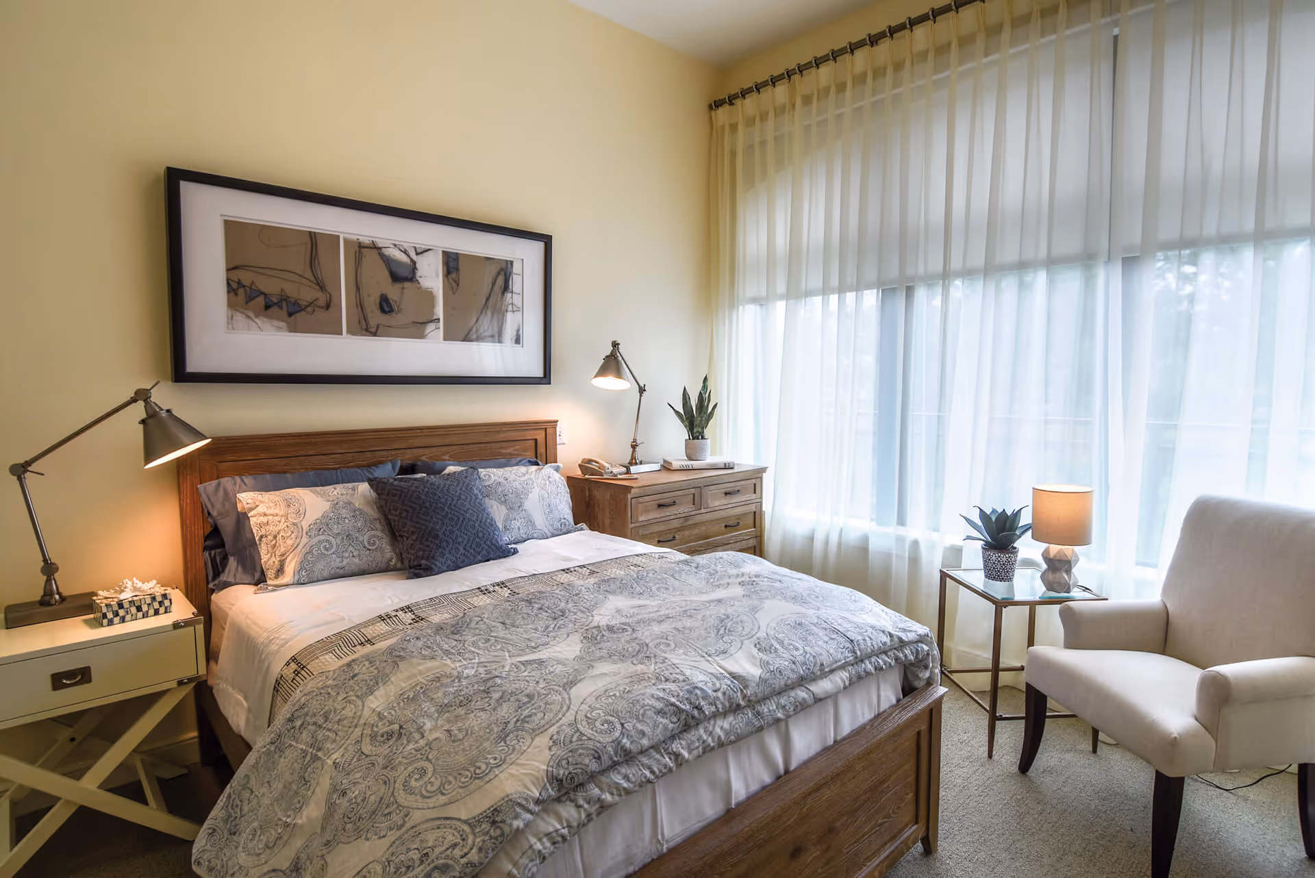 A cozy bedroom with a wooden bed frame, patterned bedding, and multiple pillows. There is a nightstand with a lamp and a tissue box on the left side of the bed. On the right side, there is a wooden dresser with a lamp, a phone, and a potted plant. A comfortable armchair and a small glass-top side table with a lamp and a potted plant are placed near a large window with sheer white curtains.